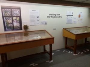 A museum exhibit with two display cases and posters on the wall between with the words "Walking with the Wordsworths." There are paper footprints on the ground creating a trail around the display cases. 