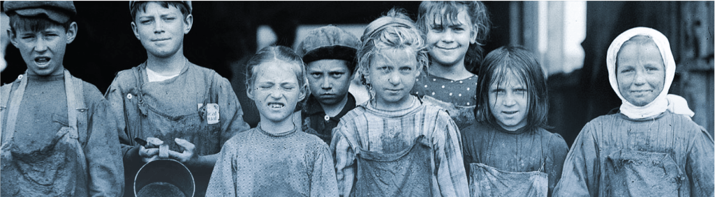 Black and white image of eight children wearing dirty clothing from the 19th century. They are all face the camera in a line, some smiling and some frowning. 