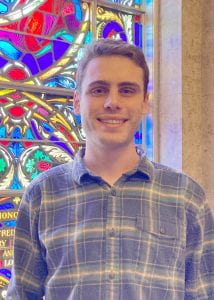 Male young adult standing in front of a stained glass window.